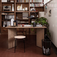 Stylish wooden desk with a minimalist black chair, surrounded by bookshelves and a green plant. Warm, earthy tones.