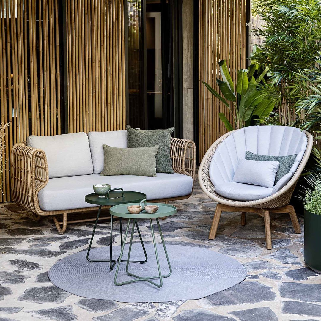 Neutral grey and white seating with green cushions, circular light grey rug, and green accent tables amidst natural greenery.