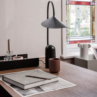 Black lamp with a conical shade, brown ceramic cup, gray notebook, and colorful stained glass window background.
