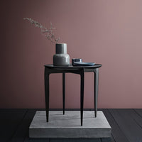 Gray side table with sleek black legs, set against a soft pink wall and a textured gray pedestal.