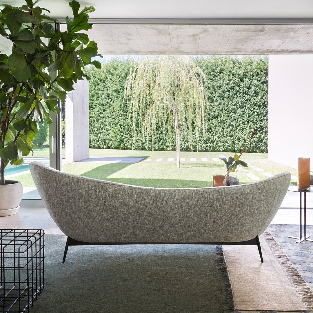 Curved light gray sofa with a textured finish, accented by green foliage and a natural outdoor backdrop.