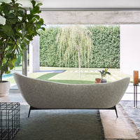 Curved light gray sofa with a textured finish, accented by green foliage and a natural outdoor backdrop.
