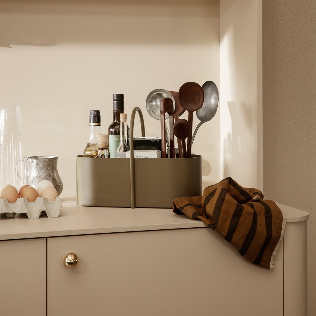 Olive green storage basket with brown and mustard striped towel, featuring kitchen tools and glassware on a beige cabinet.