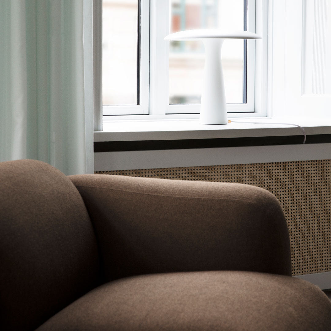 Brown upholstered chair with a smooth texture, paired with a white lamp and soft pastel curtains in the background.