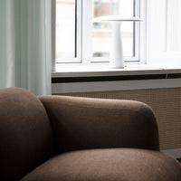 Brown upholstered chair with a smooth texture, paired with a white lamp and soft pastel curtains in the background.