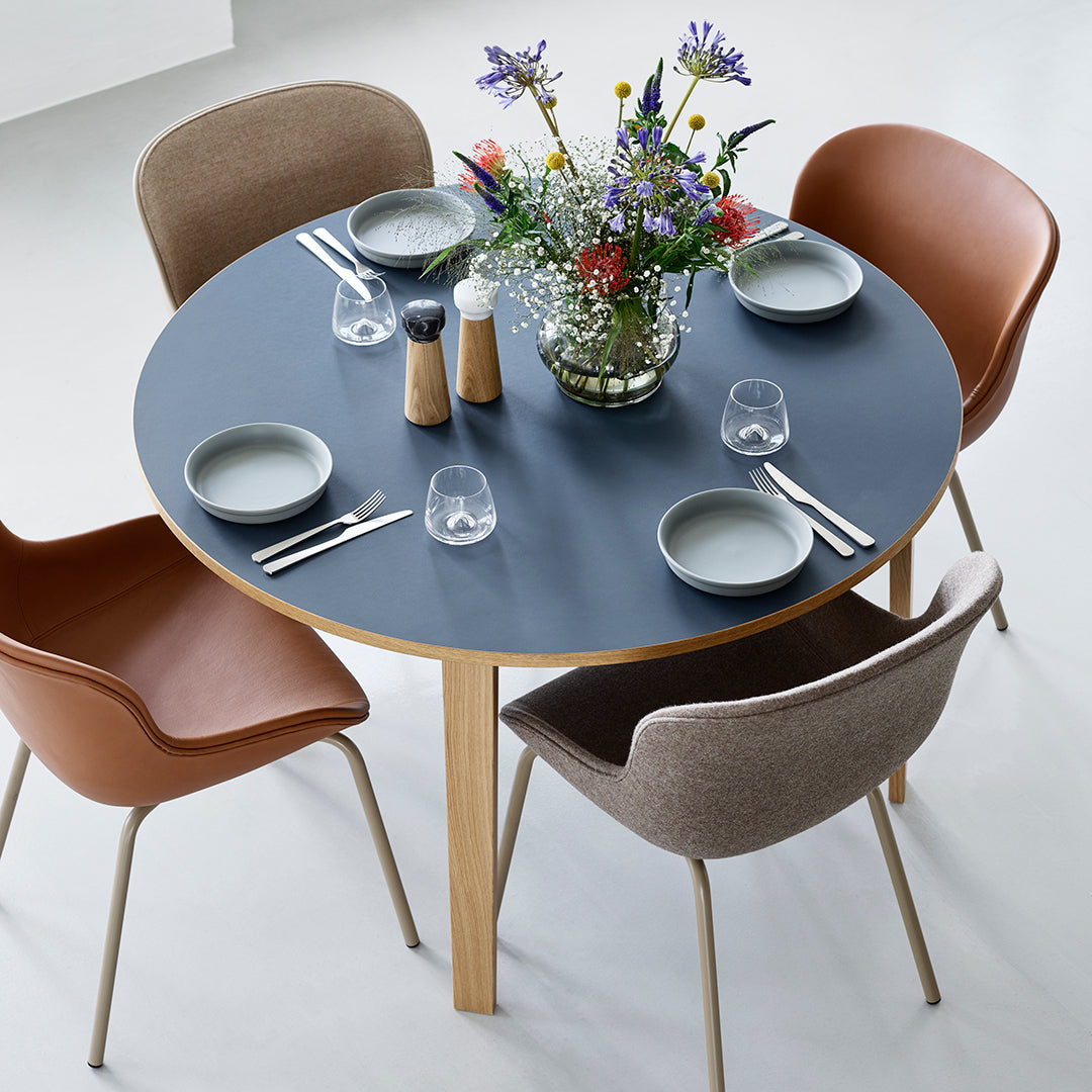 Round blue tabletop with natural wood legs, surrounded by brown and gray patterned chairs, adorned with colorful flowers.