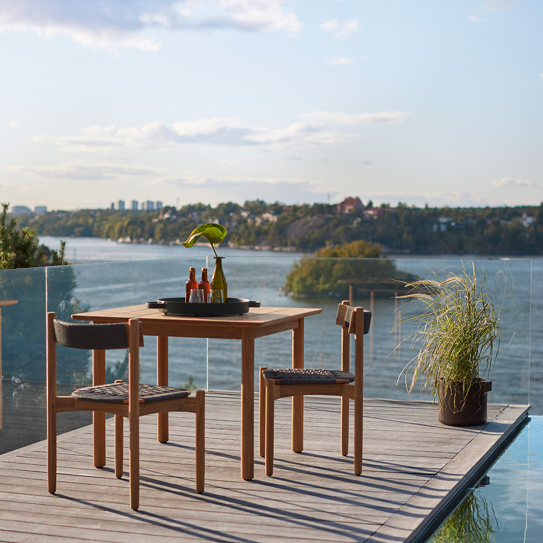 Natural wood dining table and chairs with woven fabric seats, set against a serene lakeside backdrop.
