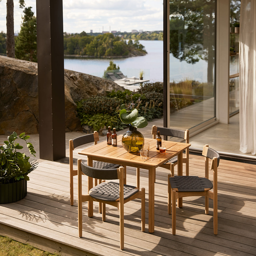 Natural wood dining table set with grey woven chairs, featuring a serene outdoor backdrop of water and greenery.