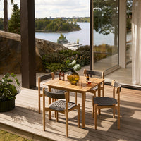Natural wood dining table set with grey woven chairs, featuring a serene outdoor backdrop of water and greenery.