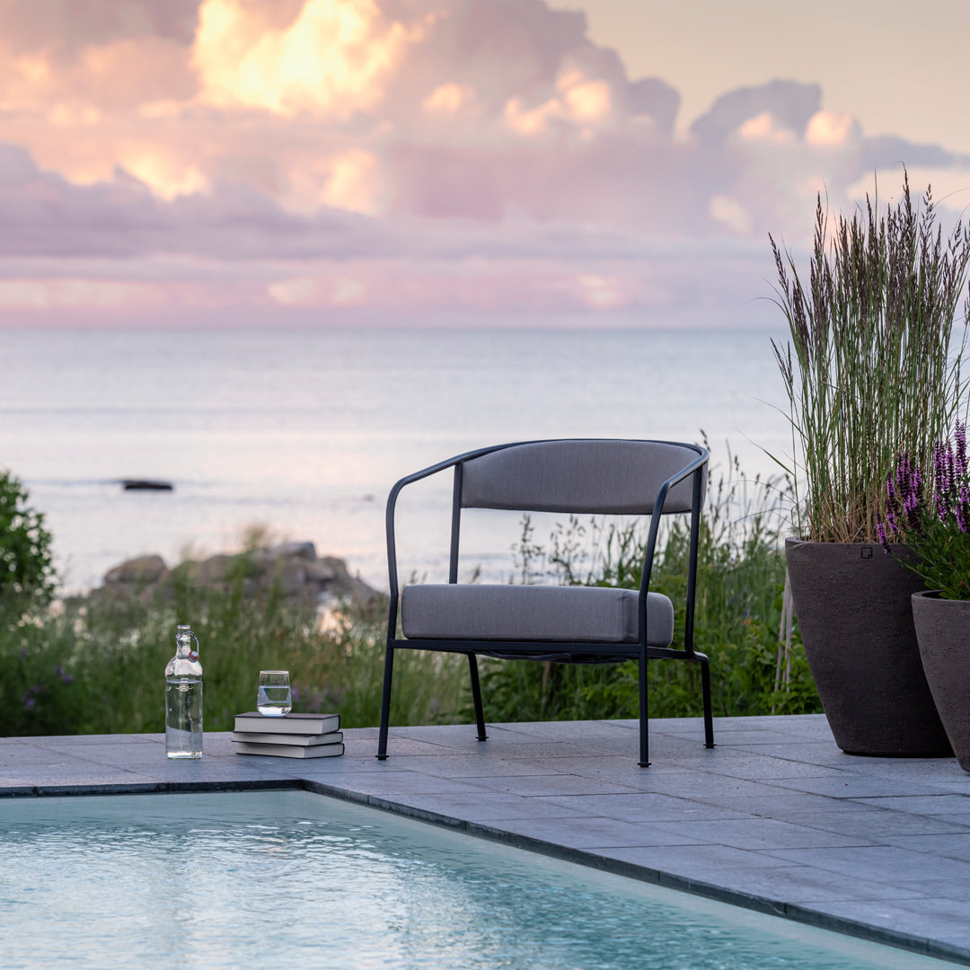 Modern black metal chair with gray cushions, set against a serene waterfront backdrop and lush greenery.