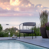 Modern black metal chair with gray cushions, set against a serene waterfront backdrop and lush greenery.