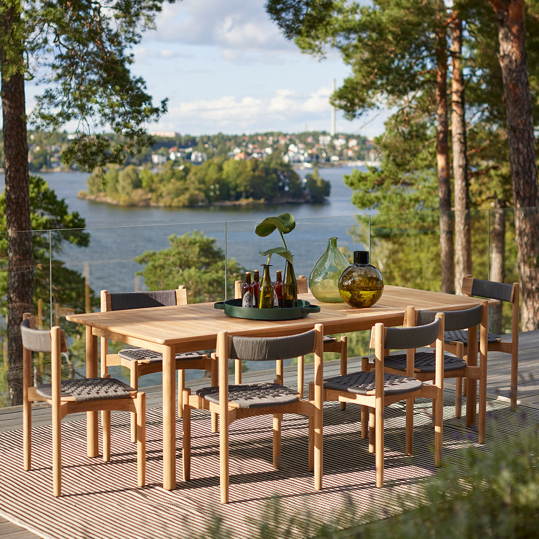 "Wooden dining table with gray patterned chairs, set against a scenic lake view and green trees."