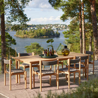 "Wooden dining table with gray patterned chairs, set against a scenic lake view and green trees."