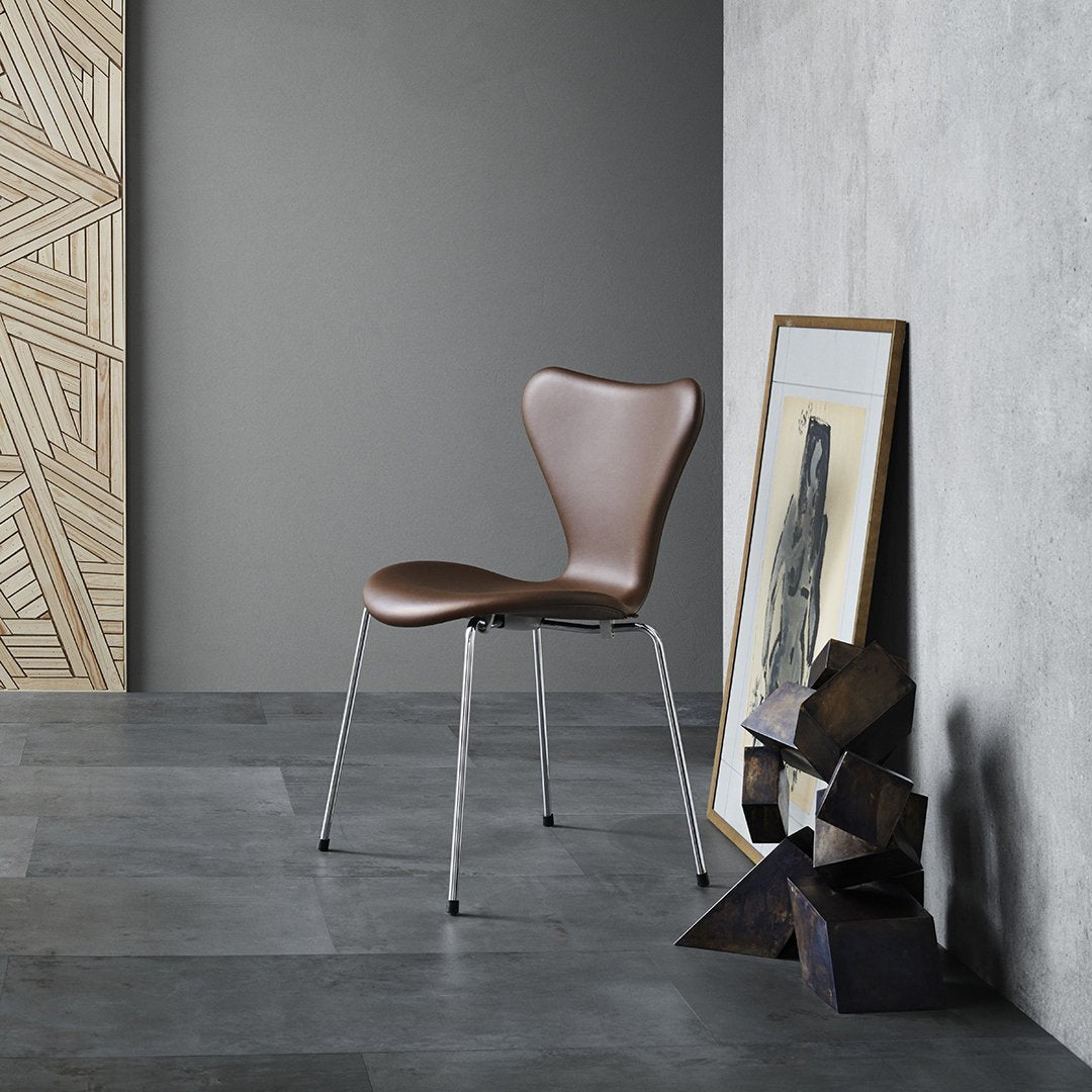Brown molded chair with sleek chrome legs, set against a textured gray wall and patterned wooden backdrop.