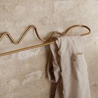 "Gold wave-shaped coat rack with a soft beige garment hanging against a textured neutral wall."