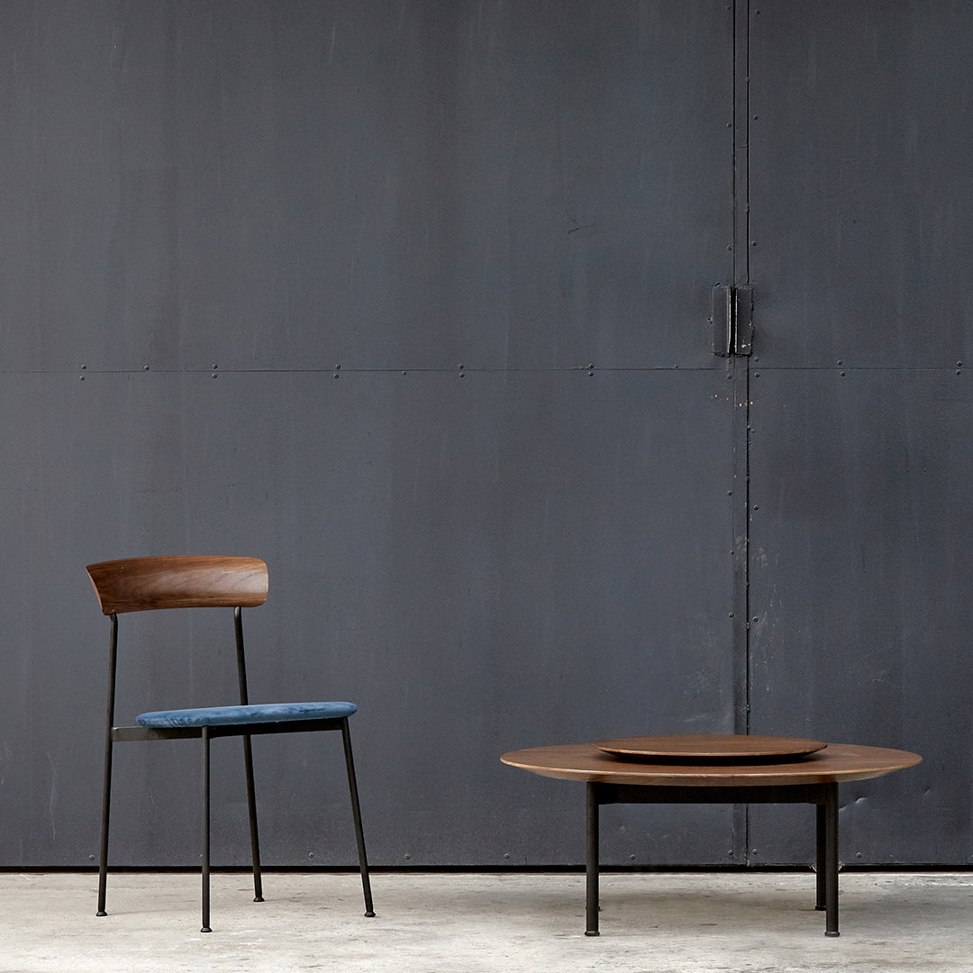 Mid-century modern chair with a dark wood back and blue seat, paired with a round wooden coffee table on a gray backdrop.