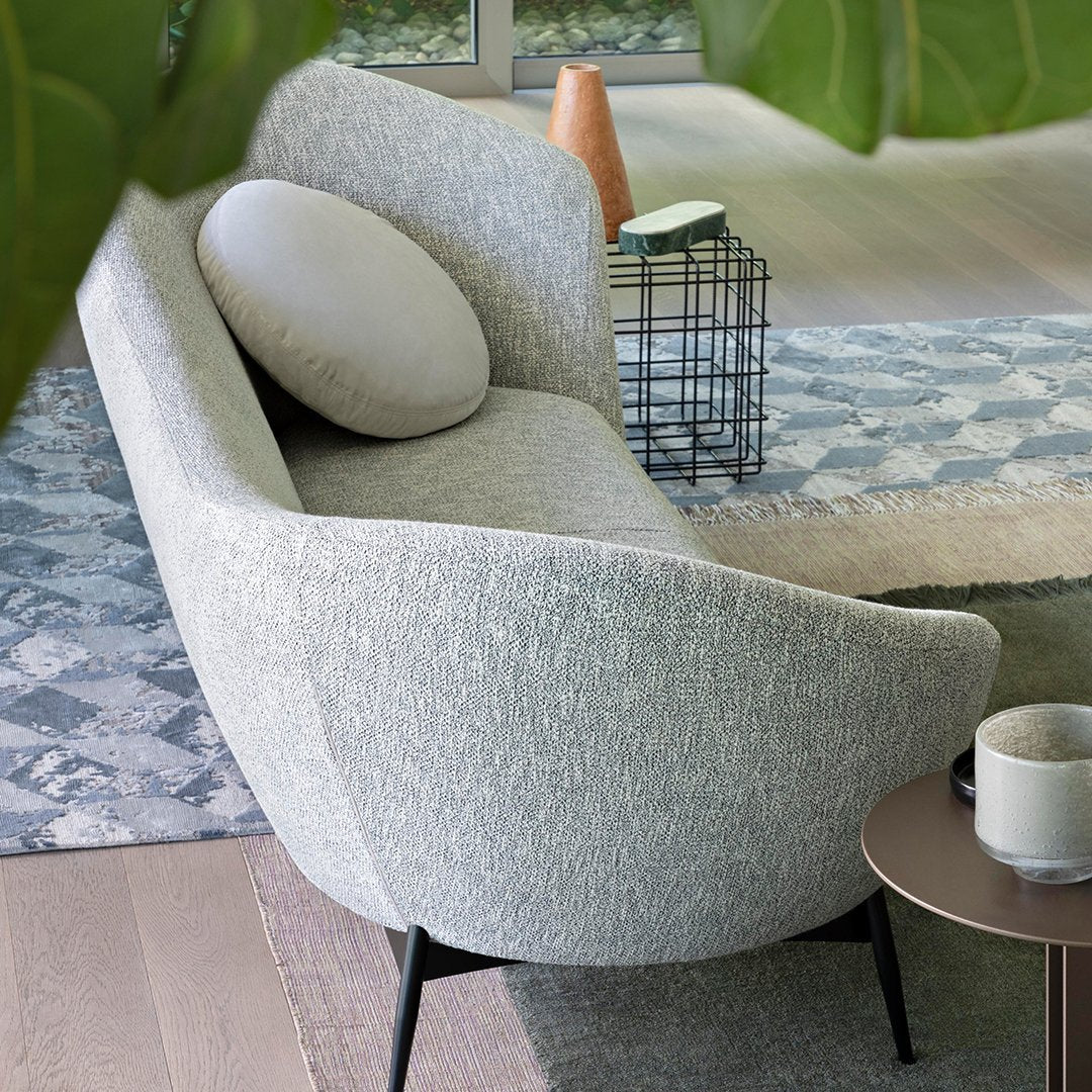 Light gray curved sofa with a round cushion, set against a patterned blue and white rug. Modern and stylish design.