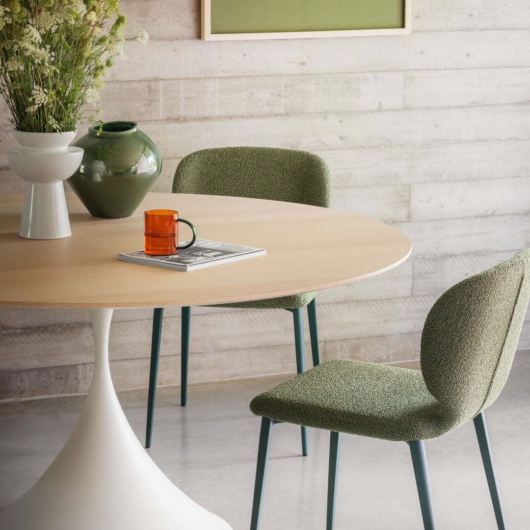 Green upholstered chairs on a round wooden table with a white base, accented by an orange mug and modern decor.