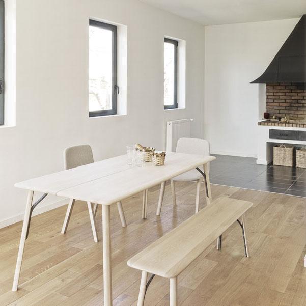 Light wooden dining table and bench in a minimalist room with white walls and dark floor, featuring simple, clean lines.