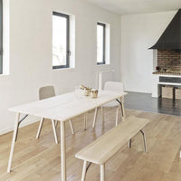 Light wooden dining table and bench in a minimalist room with white walls and dark floor, featuring simple, clean lines.