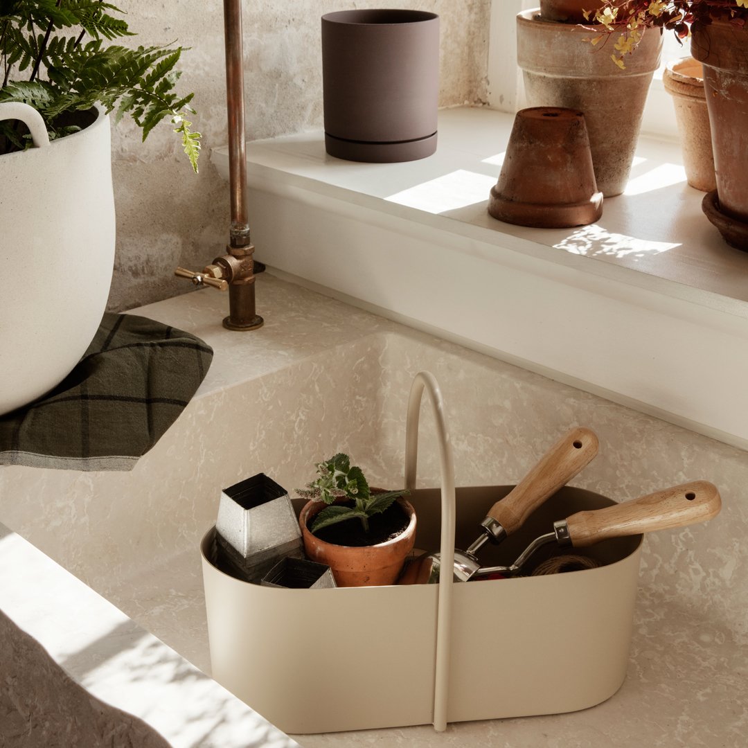 Beige gardening tool caddy with wooden handles, surrounded by green plants and earthy terracotta pots. Warm, natural tones.