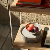 Gray plates stacked beside a bowl of red and purple fruits on a wooden shelf, with warm sunlight casting soft shadows.
