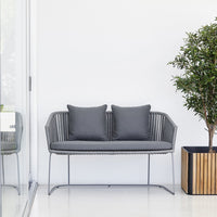 Gray outdoor sofa with two matching cushions, set against a minimalist white background and green plant accents.
