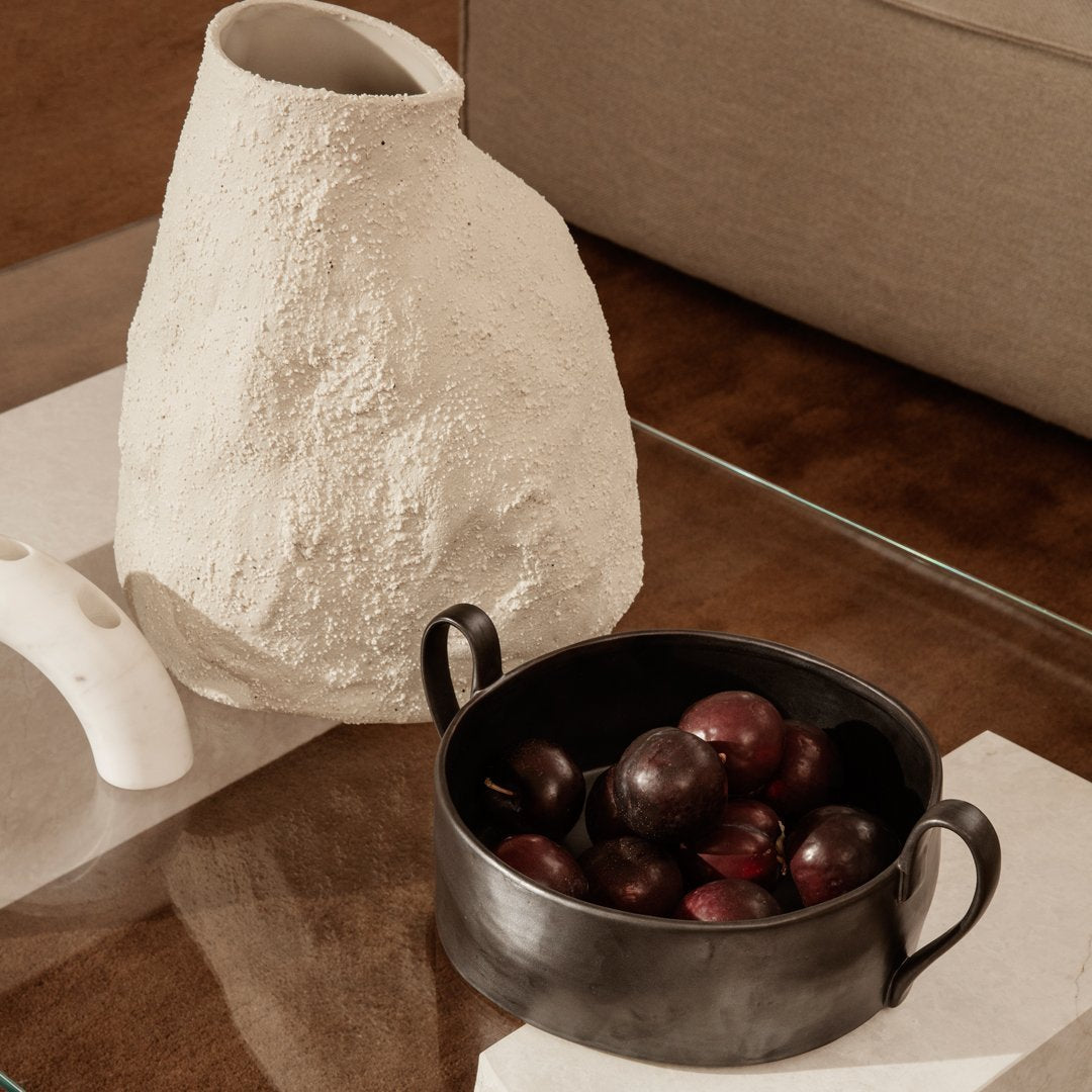 Textured white vase and black bowl filled with deep purple fruits, set on a glass table with warm brown tones.