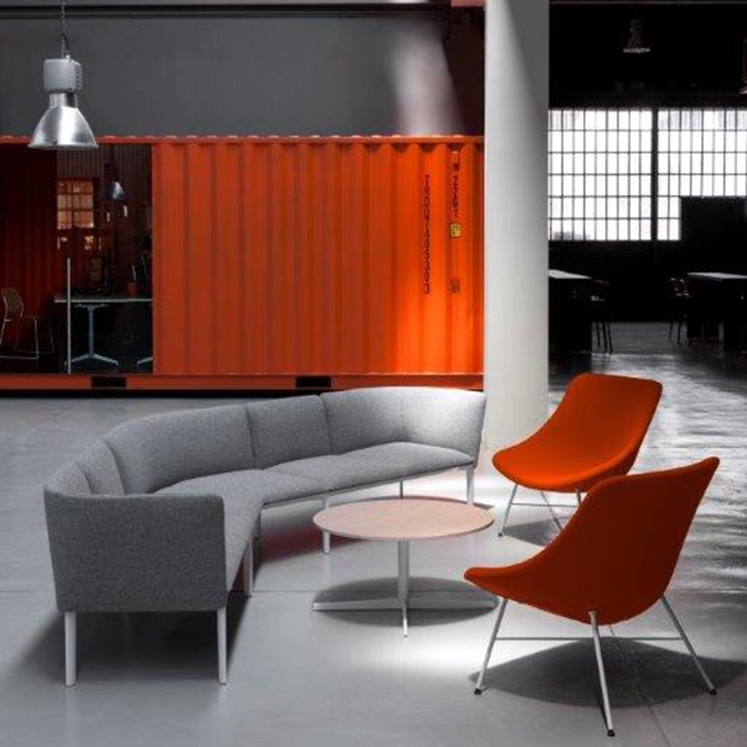 Modern gray curved sofa with vibrant orange chairs and a light wood coffee table, set against an industrial backdrop.