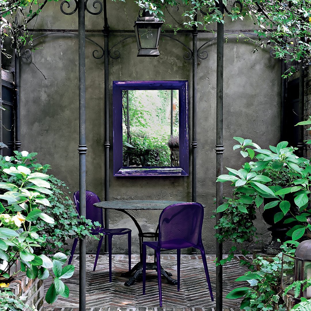 Purple chairs and mirror set against a textured grey wall, surrounded by lush green plants and brick flooring.