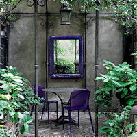 Purple chairs and mirror set against a textured grey wall, surrounded by lush green plants and brick flooring.