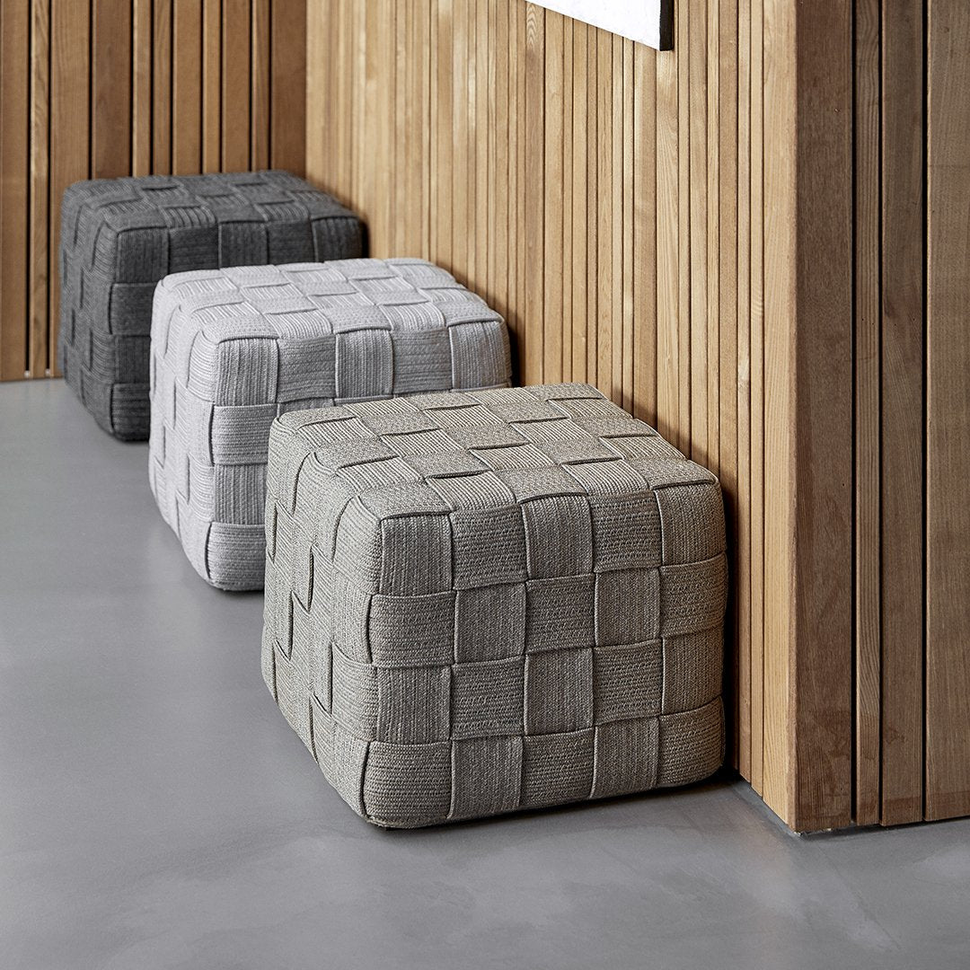 Textured square poufs in light gray, medium gray, and dark gray with woven patterns against a wooden wall.