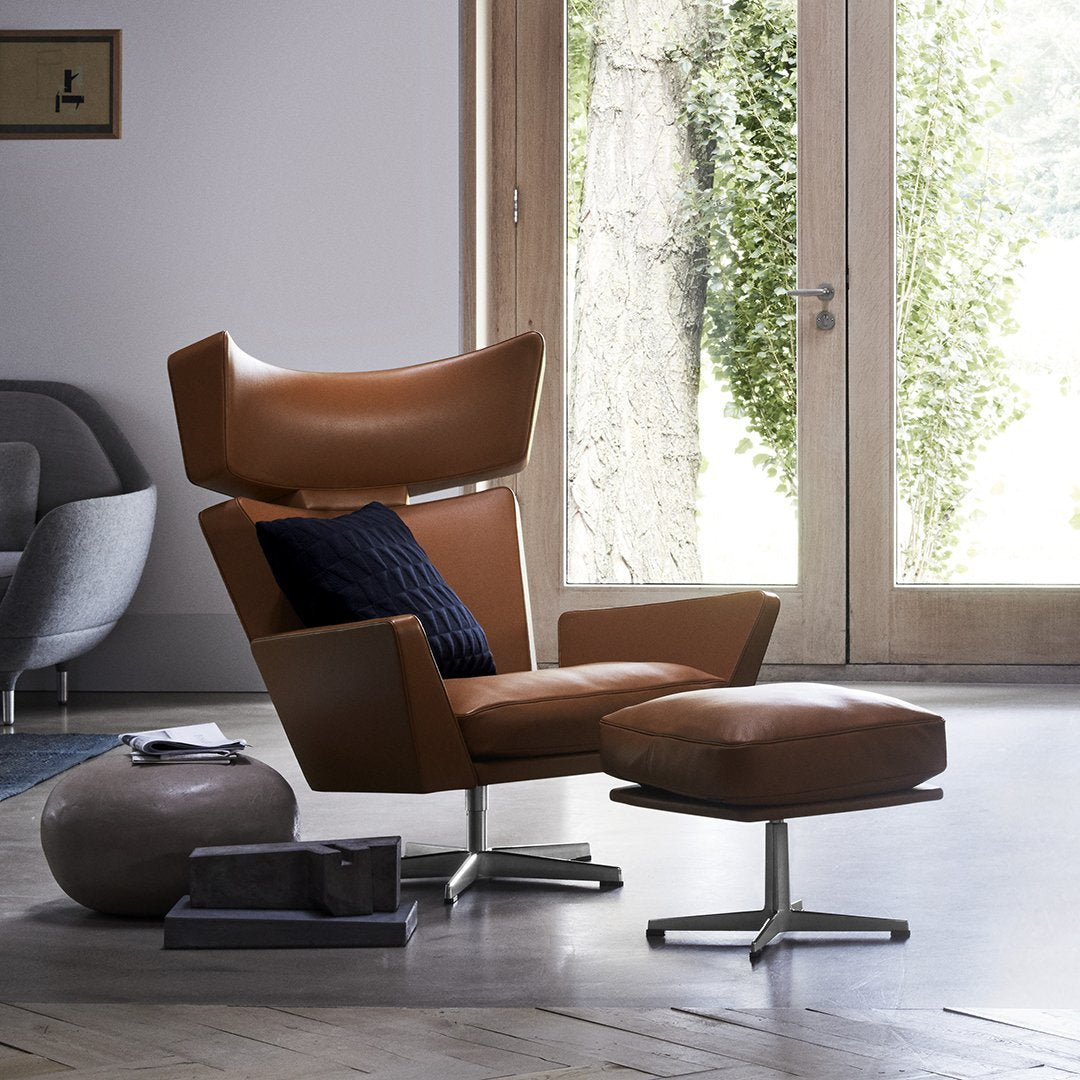 Brown leather accent chair with an oversized back, paired with a matching ottoman and a navy blue cushion. Modern design.