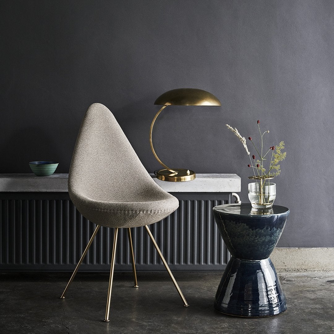 Beige textured chair with gold accents, navy ceramic stool, and muted green vase against a dark gray wall.