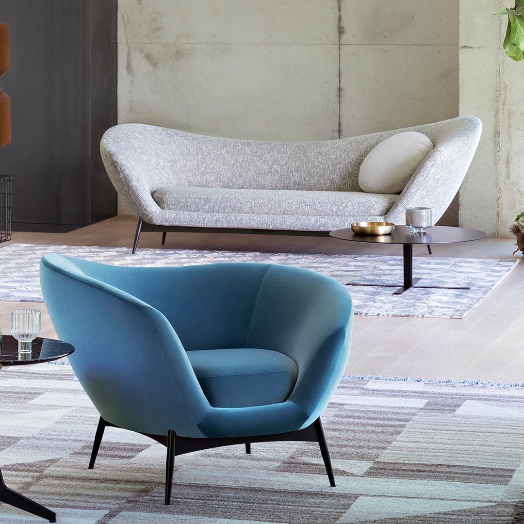 Modern gray fabric sofa and curved blue armchair on a patterned beige rug, accented by black legs.