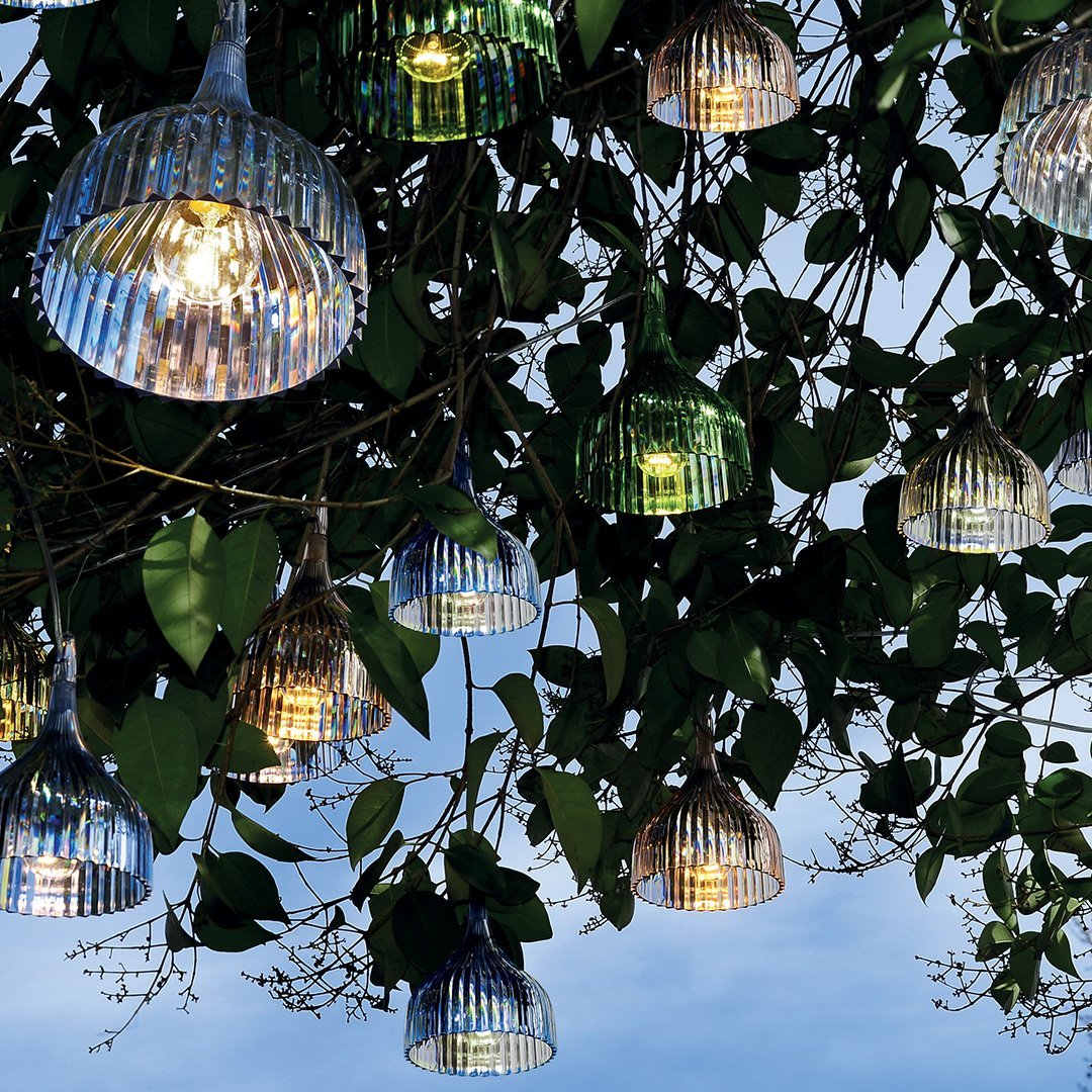 Colorful glass pendant lights with vertical stripes, hanging among lush green leaves against a twilight sky.