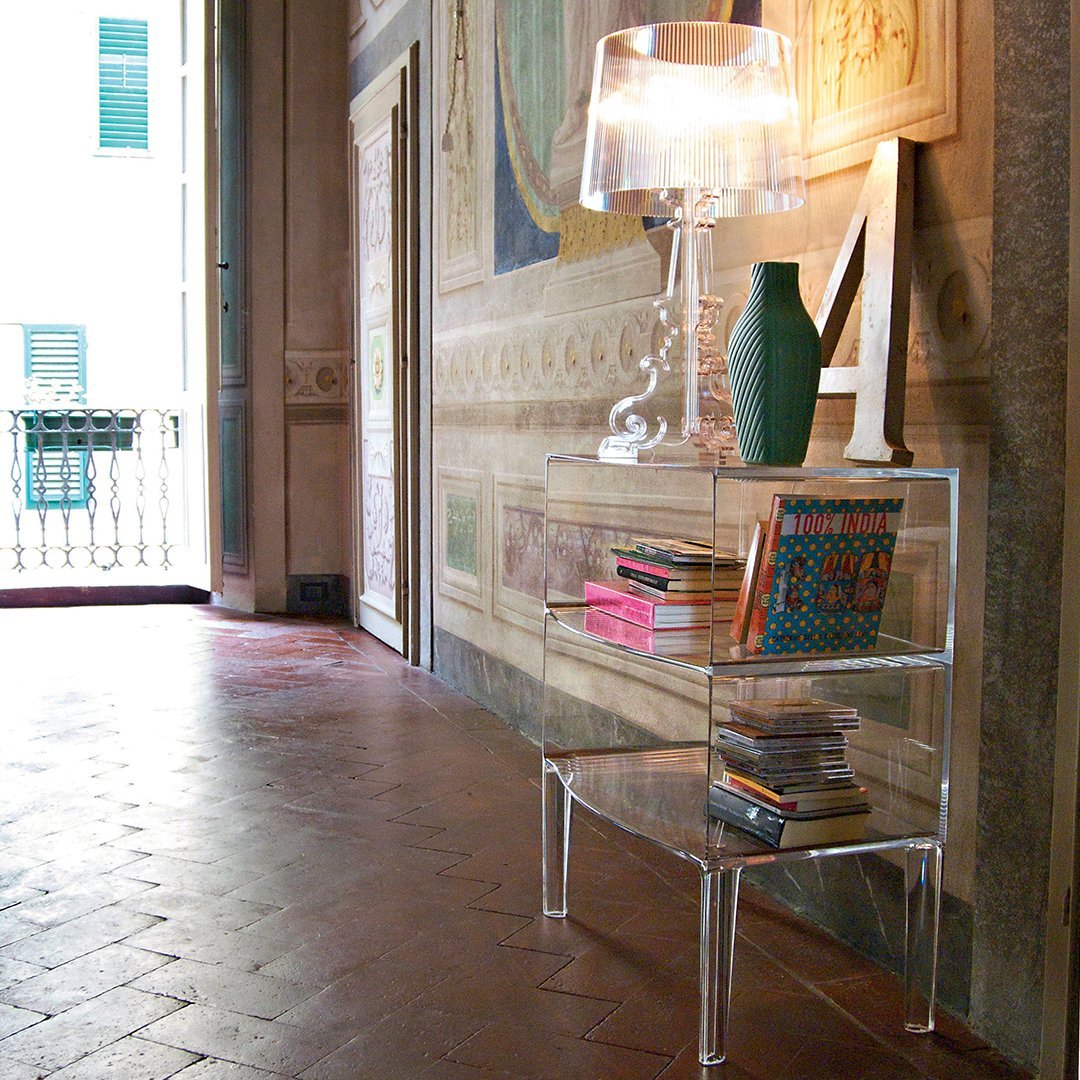 Transparent acrylic console table with colorful books and a green vase, set against a classic patterned wall.