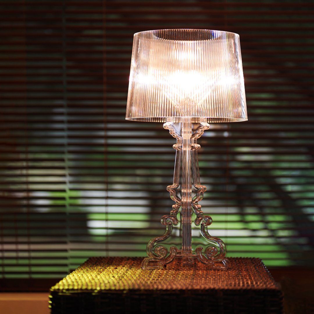 Clear acrylic lamp with a textured shade illuminating a warm, cozy ambiance. Blinds create soft shadow patterns.