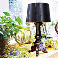 Black pleated lampshade on a glossy black base, surrounded by vibrant green plants and cacti.