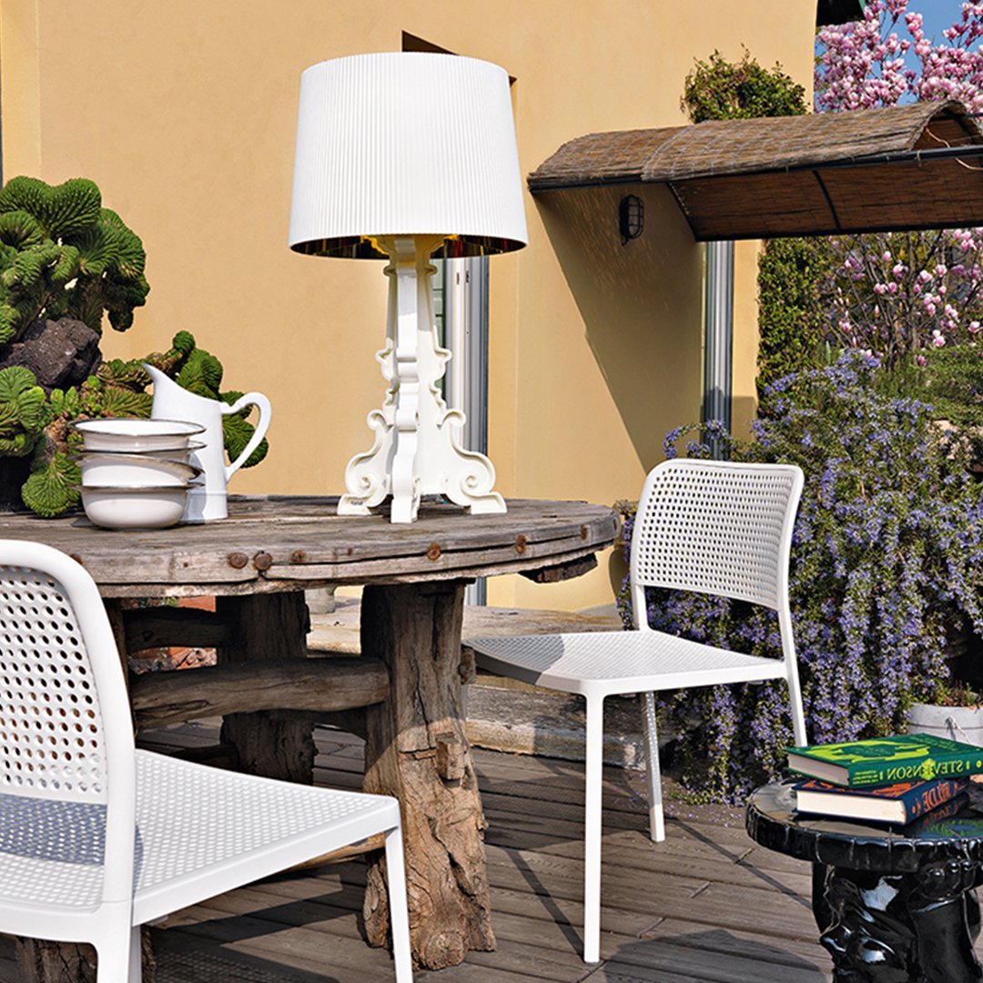 White dining chairs and lamp, natural wooden table, lush greenery, and purple wisteria flowers create a vibrant outdoor setting.