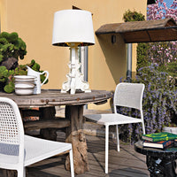 White dining chairs and lamp, natural wooden table, lush greenery, and purple wisteria flowers create a vibrant outdoor setting.