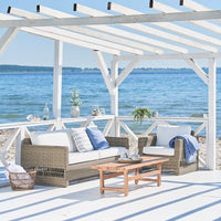 Cozy outdoor seating with white cushions, blue accents, and natural wicker under a sunlit pergola by the water.