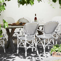 White outdoor chairs with black woven patterns arranged around a rustic wooden table in a lush garden setting.