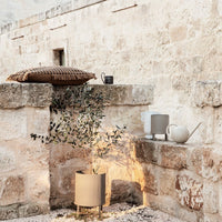 Decorative earthy tones and textures, featuring a woven pillow, ceramic pots, and greenery against a stone backdrop.