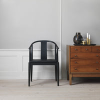 Black modern chair with a sleek design, set against a neutral gray wall and accompanied by a wooden dresser.