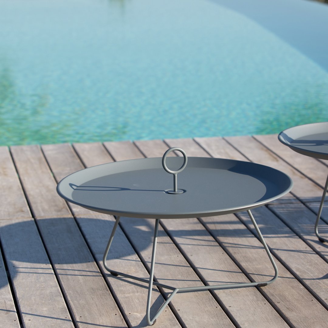 Gray round coffee table with minimalistic design and sleek metal base, set against a tranquil poolside backdrop.
