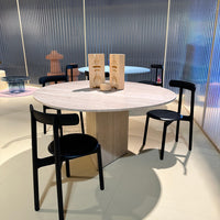 Modern round dining table with a light wood finish, surrounded by sleek black chairs against a pastel backdrop.