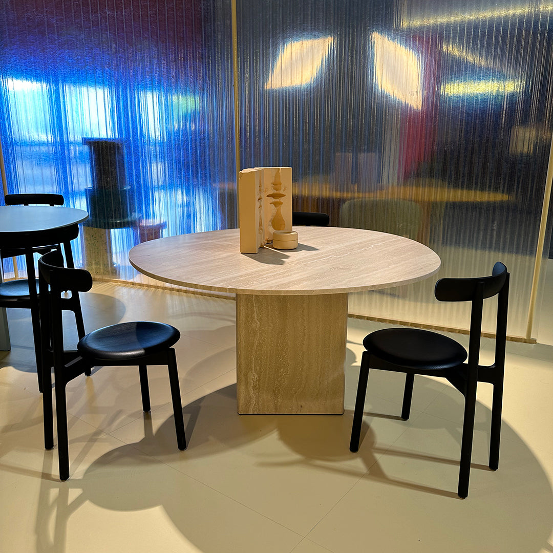 Round wooden table with a light wood finish, accompanied by two sleek black chairs against a colorful backdrop.