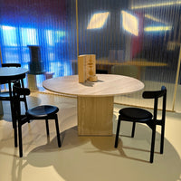 Round wooden table with a light wood finish, accompanied by two sleek black chairs against a colorful backdrop.