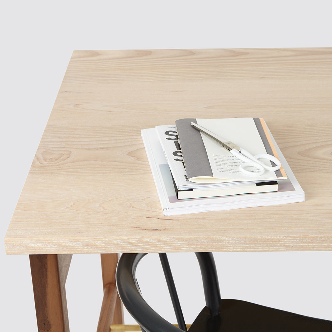 Light wood desk with a smooth finish, featuring a white and gray stationery stack and a black chair.